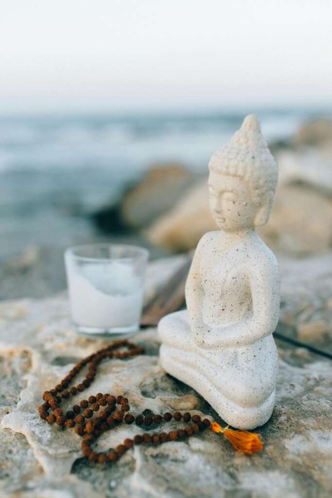 Peaceful Buddha figurine on rocks by the ocean, accompanied by meditation beads and a candle.